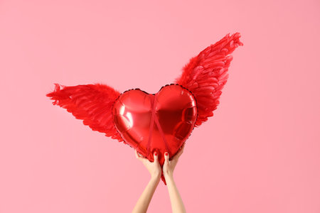Female hands holding heart-shaped balloon with Cupid's wings on pink background. Valentine's Day celebrationの写真素材