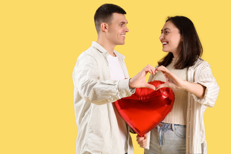 Young couple with balloon making heart gesture on yellow background. Valentine's Day celebration