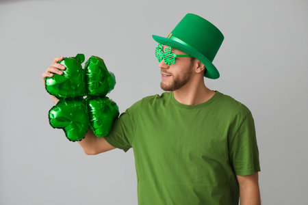Young man in leprechaun hat and decorative glasses with air balloon in shape of clover on gray...