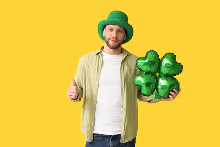Young man in leprechaun hat with air balloon in shape of clover showing thumb-up gesture on yellow background. St. Patrick's Day celebrationの写真素材