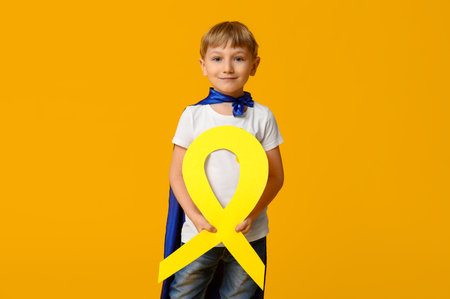 Cute little boy in superhero costume with yellow ribbon on color background. International Childhood Cancer Dayの写真素材