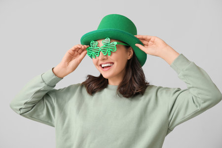 Beautiful young woman in leprechaun hat and decorative glasses in shape of clover on gray background. St. Patrick's Day celebrationの写真素材