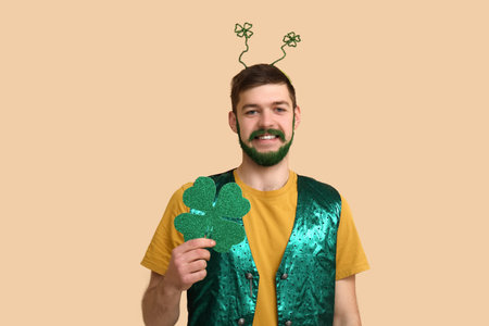 Young man in headband with green beard holding clover on beige background. St. Patrick's Day...