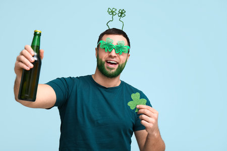 Young man in headband with green beard holding bottle of beer and clover on blue background. St. Patrick's Day celebrationの写真素材