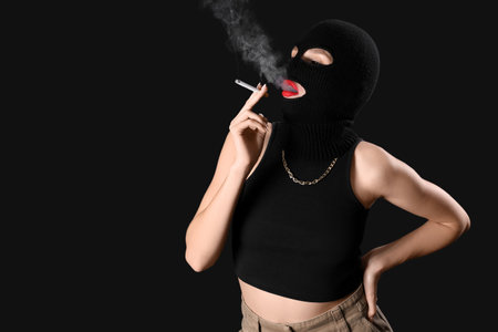 Beautiful young woman in balaclava smoking cigarette on black backgroundの写真素材