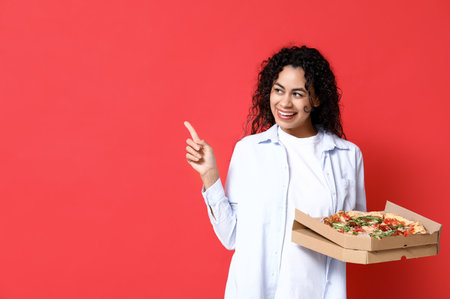 Beautiful young happy African-American woman with delicious pizza pointing at something on red backgroundの写真素材