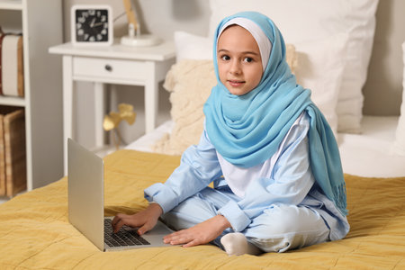 Little Muslim girl in hijab sitting on bed with modern laptop at homeの写真素材