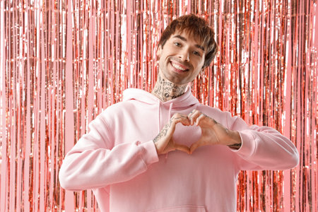 Happy young man making heart with hands and tinsel curtain on pink background. Valentine's day celebrationの写真素材