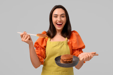 Happy young woman holding frying pan with tasty pancakes and spatula on gray backgroundの写真素材