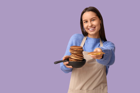 Beautiful young woman holding frying pan and spatula with tasty pancakes on lilac backgroundの写真素材