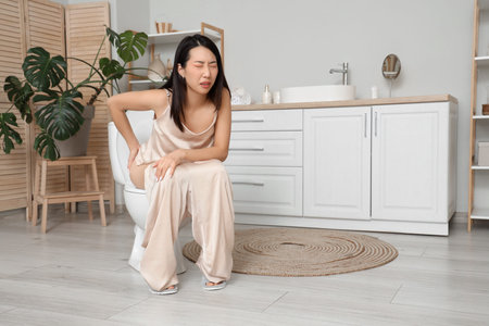 Young Asian woman with hemorrhoids sitting on toilet bowl in restroomの写真素材