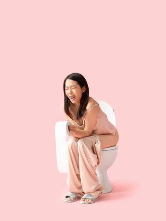 Young Asian woman in pajamas with hemorrhoids sitting on toilet bowl against pink backgroundの写真素材