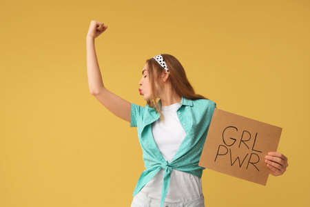 Happy young pin-up woman holding card with text GIRL POWER and kissing her muscles on yellow backgroundの写真素材