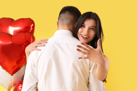 Young engaged couple with wedding ring and heart-shaped balloons hugging on yellow background. Valentine's Day celebrationの写真素材