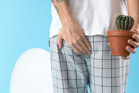 Young man with cactus and toilet bowl on blue background, back view. Hemorrhoids concept