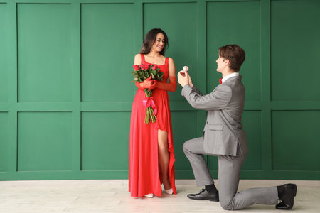 Young man with ring proposing to his girlfriend near green wall. Valentine's Day celebrationの写真素材