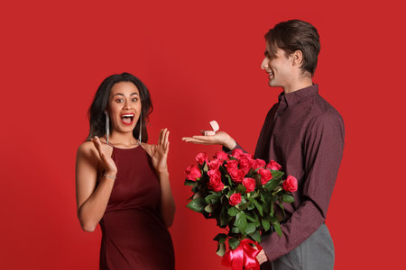 Young man with roses and ring proposing to his girlfriend on red background. Valentine's Day celebrationの写真素材