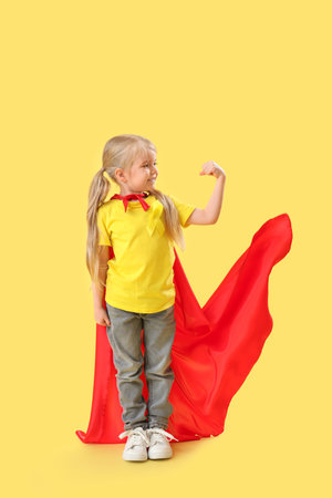 Cute little girl in superhero costume with yellow ribbon showing muscles on color background. Childhood cancer awareness conceptの写真素材