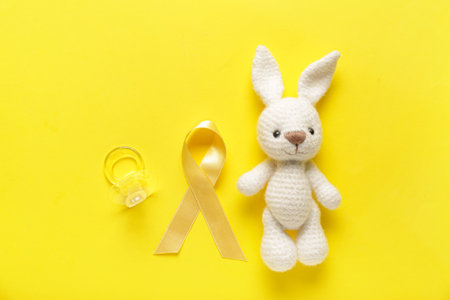 Toy bunny with golden ribbon and pacifier on yellow background. Childhood cancer awareness conceptの写真素材