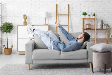 Handsome young man with air conditioner remote control lying on sofa in living roomの写真素材