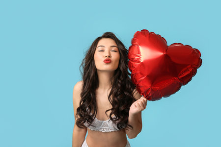Beautiful young Asian woman in sexy underwear with heart-shaped balloon blowing kiss on blue backgroundの写真素材