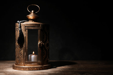 Muslim lamp with burning candle and prayer beads for Ramadan on wooden table against dark background