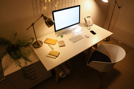 Blank computer monitor with stationery and glowing lamps in office at nightの写真素材