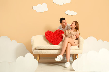 Young couple with heart sitting on sofa in clouds near beige wallの写真素材