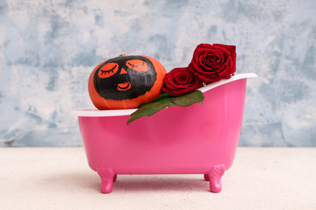 Pumpkin with drawn face, clay mask and roses in miniature bathtub on white table