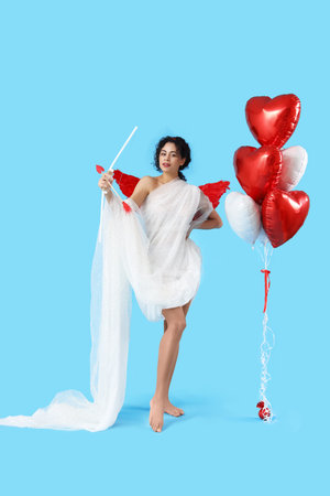 Young African-American woman dressed as Cupid with bow and balloons on blue background. Valentine's...