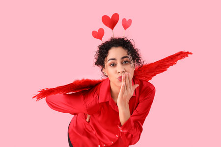 Young African-American woman with Cupid's wings and hearts blowing kiss on pink background....