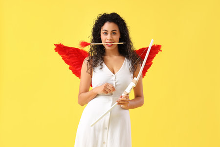 Young African-American woman dressed as Cupid with bow and arrow on yellow background. Valentine's Day celebrationの写真素材