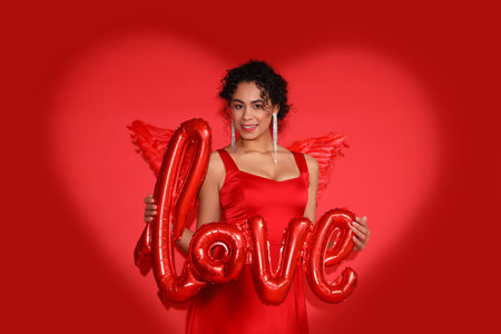 Young African-American woman dressed as Cupid with the word LOVE made of balloons on red background....
