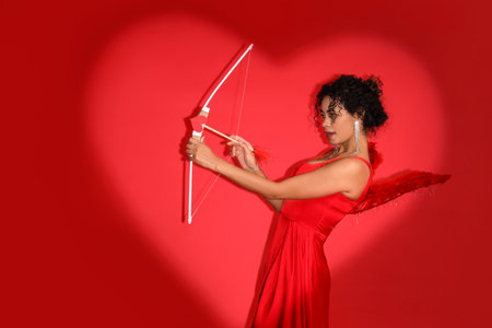 Young African-American woman dressed as Cupid with bow on red background. Valentine's Day celebrationの写真素材
