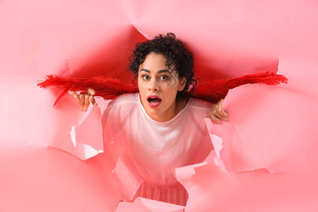 Shocked young African-American woman dressed as Cupid visible through torn pink paper. Valentine's...