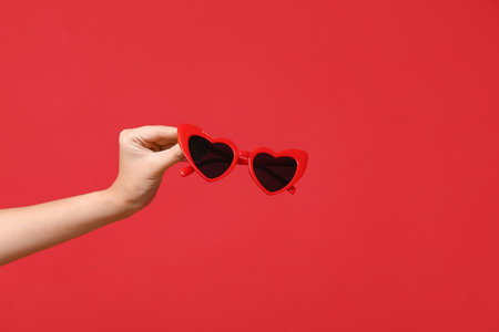 Female hand holding heart-shaped sunglasses on red background. Valentine's Day celebrationの写真素材