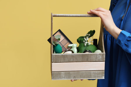 Female hand with wooden basket full of decorative cosmetics and Easter bunnies on yellow backgroundの写真素材