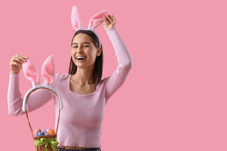Young woman in bunny ears with basket of Easter eggs on pink background