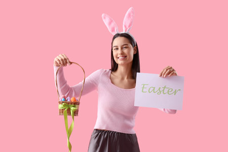 Young woman in bunny ears holding paper with the word EASTER and basket of eggs on pink backgroundの写真素材