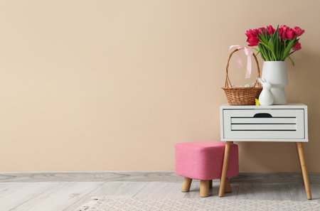 Vase with tulips and basket of Easter eggs on table near beige wall in roomの写真素材