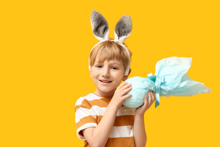 Cute little boy in bunny ears with Easter gift egg on yellow background