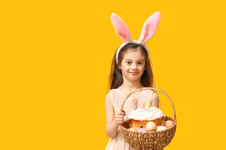 Cute little girl in bunny ears with basket of Easter cake and eggs on yellow background