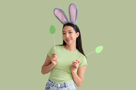 Young Asian woman in bunny ears with paper Easter eggs on green background