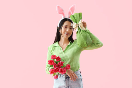 Young Asian woman in bunny ears with tulips Easter gift egg on pink backgroundの写真素材