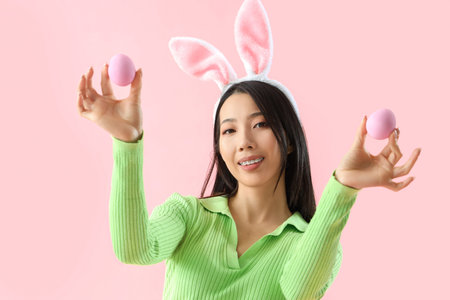 Young Asian woman in bunny ears with Easter eggs on pink background