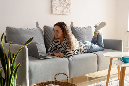 Young woman in eyeglasses using laptop on gray sofa at homeの写真素材