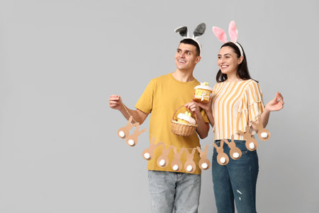 Happy young couple with Easter cakes, garland and bunny ears on gray background