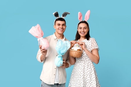 Happy young couple with Easter bunny ears, basket and gifts on blue background