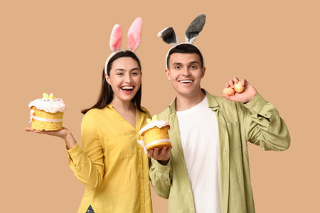Happy young couple with Easter cakes, eggs and bunny ears on beige backgroundの写真素材