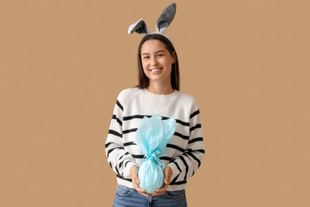 Pretty young woman with bunny ears and Easter gift on beige background